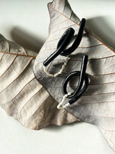Load image into Gallery viewer, Close-up of Pearl Twist Earrings featuring twisted black leather loops paired with a strand of white pearls. The sculptural, lightweight design is bold and unusual, handcrafted from sustainable materials to create a modern statement piece."