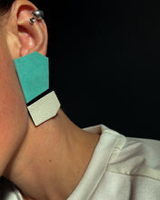 Load image into Gallery viewer, Close-up of a person wearing a geometric leather earring in turquoise, black, and white. The earring has bold, angular shapes with contrasting textures, creating a modern, structured look against a dark background.