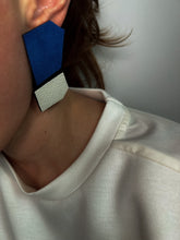 Load image into Gallery viewer, Close-up of a person wearing geometric leather earrings in blue, black, and white. The earrings feature bold angular shapes with contrasting textures.