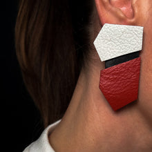 Load image into Gallery viewer, Close-up of a person wearing geometric leather earrings in red, black, and white. The earrings feature bold angular shapes with contrasting colours.