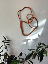 Load image into Gallery viewer, Rust-colored abstract wall sculpture on a white wall with green leaves in the foreground.