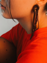 Load image into Gallery viewer, Close-up of a person wearing brown long beaded leather  earrings and a red garment with a neutral background