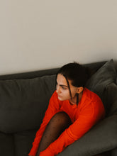 Load image into Gallery viewer, Woman in an orange sweater and long brown leather earringssitting on a dark gray couch against a plain wall.