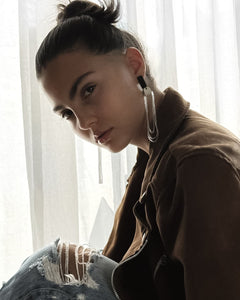 Person wearing a brown jacket and ripped jeans sitting in front of white curtains wearing a dangling leather glass bead earring 