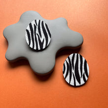 Load image into Gallery viewer, Close-up of earrings featuring a white leather base with bold, hand-cut black zebra stripes. The modern design showcases sharp contrast and wild energy, blending edgy patterns with effortless style. Lightweight and handcrafted from sustainable materials, these earrings are part of a nature-inspired collection.