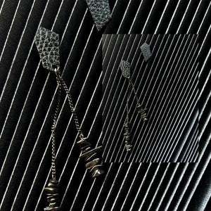 a pair of earrings designed for individuals seeking a unique and edgy accessory. The earrings are mismatched studs made from leather with a geometric shape. One earring features a simple stud design, while the other earring has a longer drop style with two black stainless chains of different lengths adorned with hematite stones.