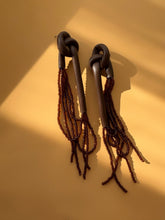Load image into Gallery viewer, A pair of handcrafted earrings with knotted espresso leather tops, cascading strands of brown glass beads, and stainless steel studs, photographed on a warm background.