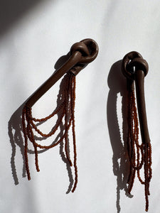 A pair of handcrafted earrings with knotted espresso leather tops, cascading strands of brown glass beads, and stainless steel studs, photographed on a white background.